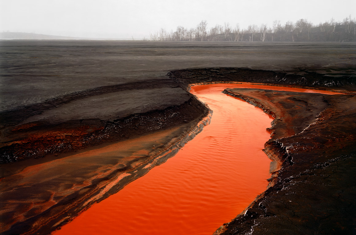 A black sandy landscape with small red river flowing through it and some dead trees in the distant background.